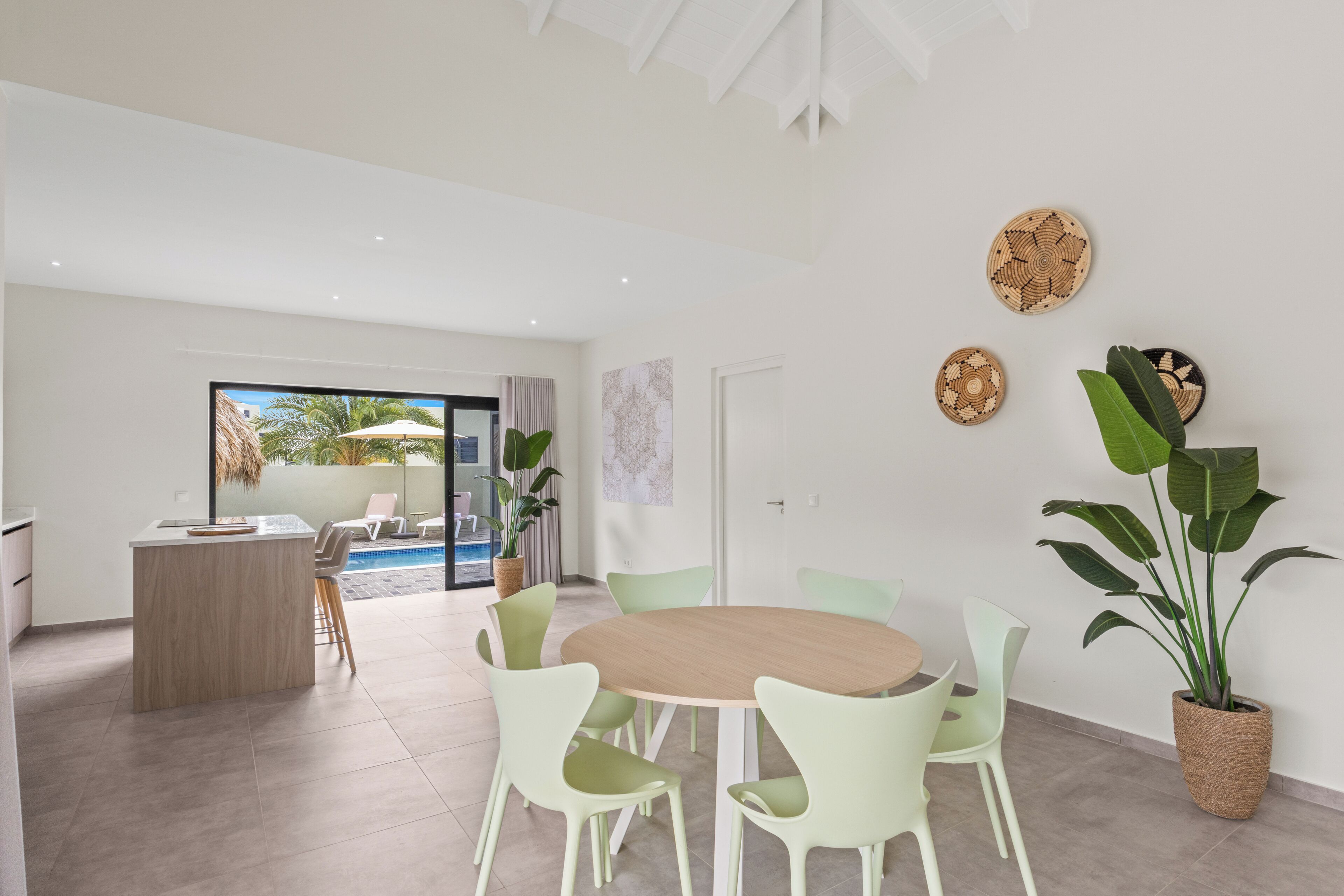 A modern living room with a round wooden table and light green chairs, a kitchen area with a bar, and a view of an outdoor pool.