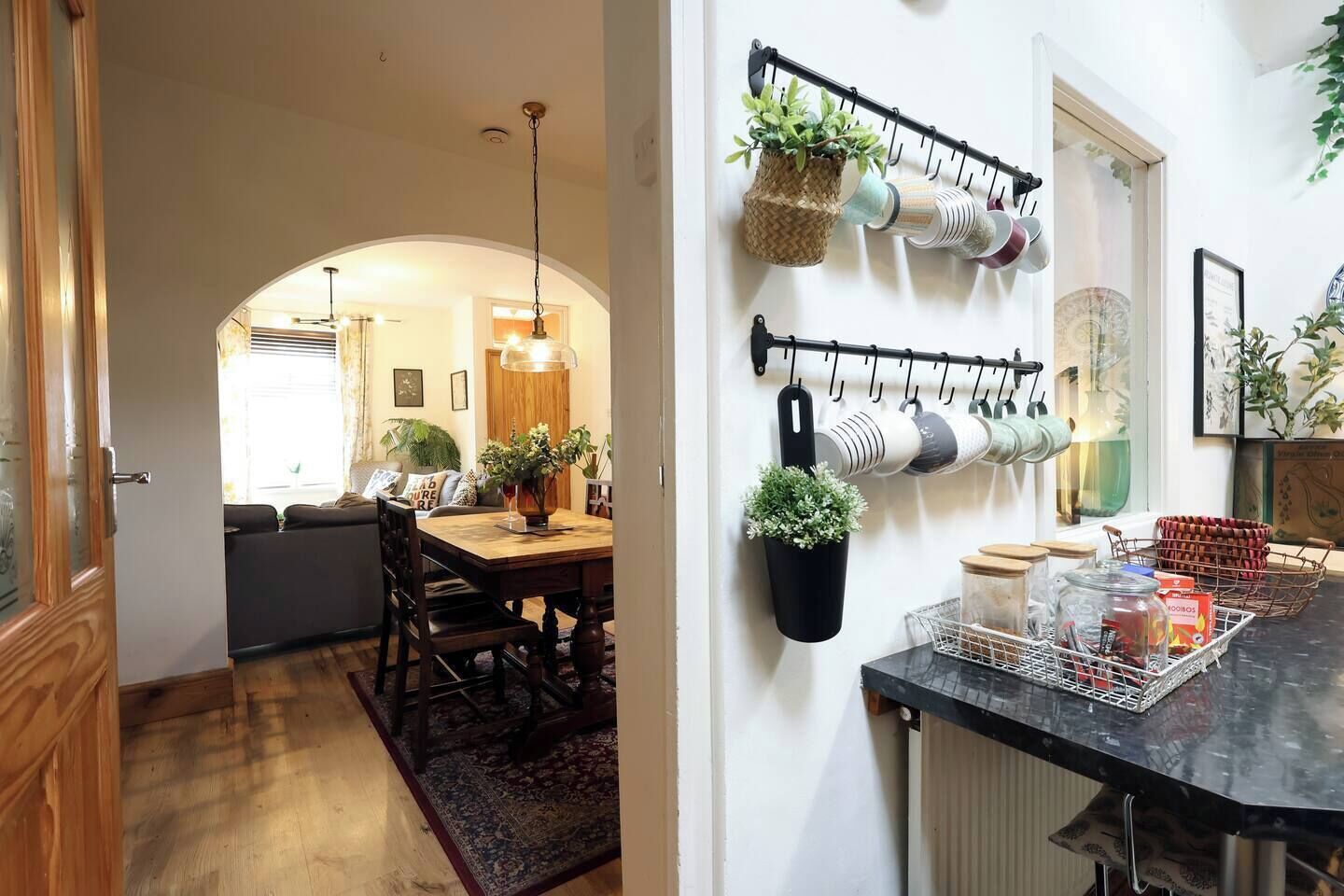 A dining area with a wooden table and chairs, a hanging plant, and a kitchen counter with a basket and jars.