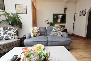 Ein Wohnzimmer mit grauem Sofa, einem gemusterten Sessel, einem Couchtisch mit Blumen und einer Fernbedienung.