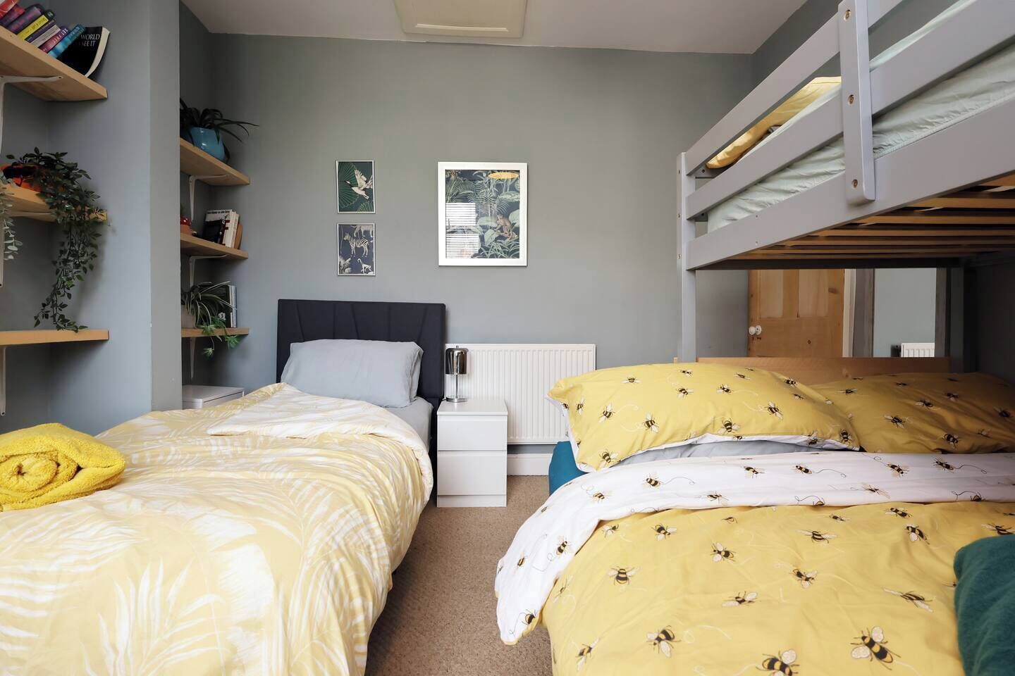 Ein Schlafzimmer mit Etagenbett, Nachttisch, Heizkörper und Wandregalen mit Büchern und Pflanzen.