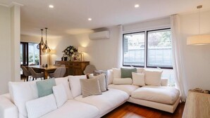 A modern living room with a sectional sofa, a dining area with a wooden table, and large windows with curtains. - 9 Anne St Diamond Cr (Diamond Creek)