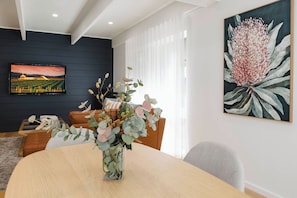 A modern living room with a dining table, a sofa, a TV mounted on the wall, and a large floral painting. - 9 Anne St Diamond Cr (Diamond Creek)