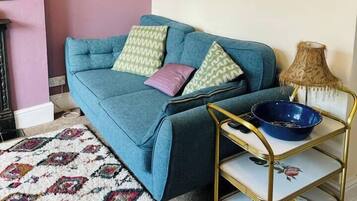 A living room with a blue sofa, patterned rug, wall-mounted pictures, a hanging plant, and a gold-framed side table with a bowl and remote control.