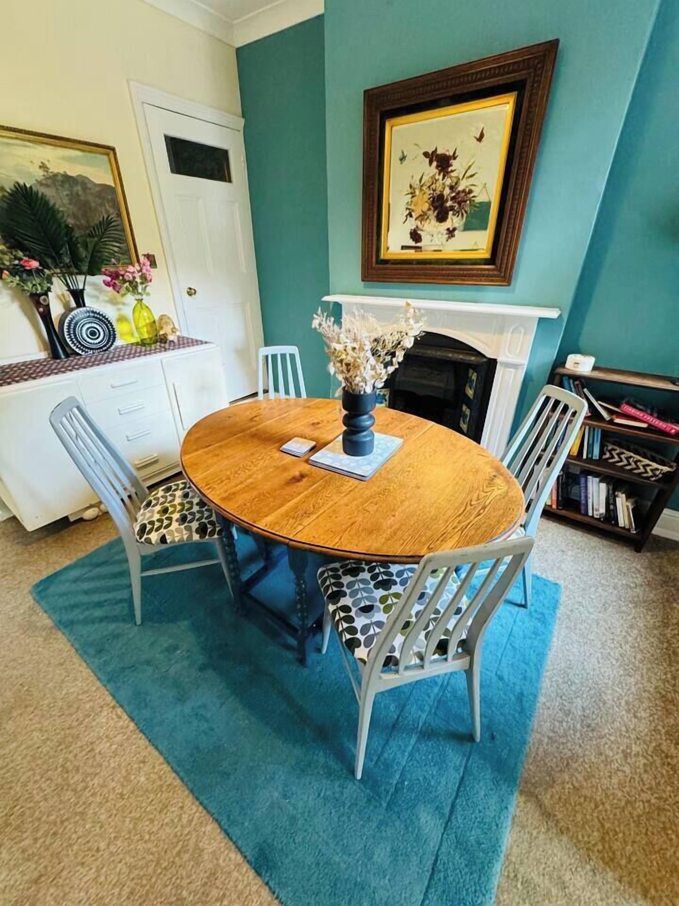 A dining area with a round wooden table, four chairs, a blue rug, a white door, a bookshelf, and a framed picture on the wall.