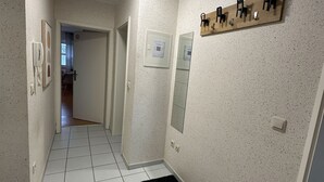 A hallway with a tiled floor, a black rug, a wooden shelf with chairs, a wall-mounted light, and a door leading to another room. - Cozy vacation rental for workers: Sleeps 4, 2 bedrooms, kitchen, bathroom, balcony (Hopsten)