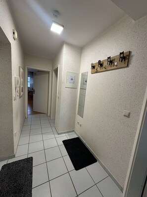 Ein Flur mit Fliesenboden, einem schwarzen Teppich, einem Holzbodenregal mit Stühlen, einer Wandleuchte und einer Tür zu einem anderen Raum.