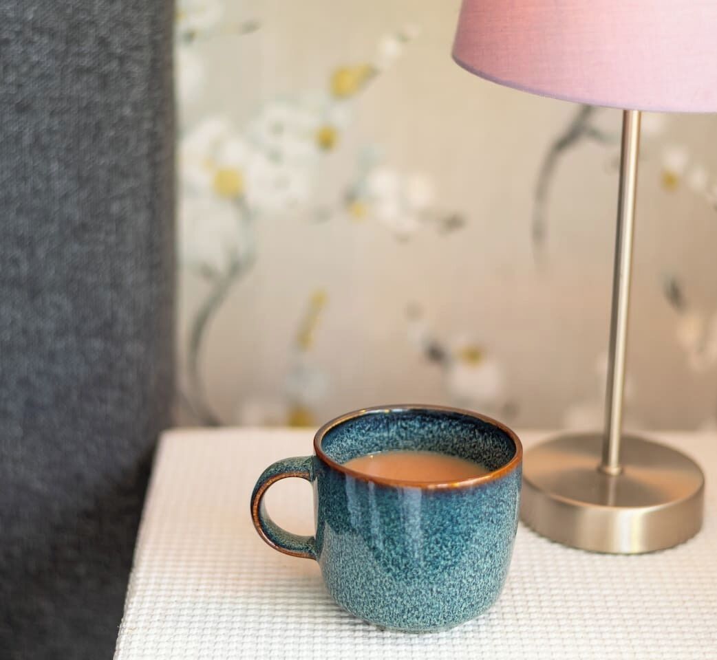 Une tasse en céramique bleue posée sur une surface blanche texturée, une lampe rose et un mur recouvert d’un papier peint à motifs floraux.