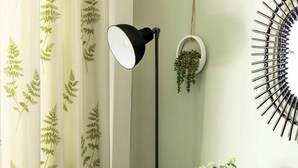 Een groene fauteuil met een bloemenkussen, een vloerlamp, een klein rond tafeltje met een vaas en een spiegel aan de muur.