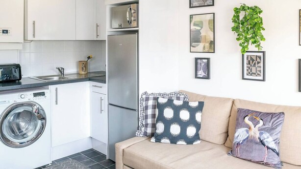 A kitchenette with a washing machine, a refrigerator, and a microwave. There is a beige sofa with patterned cushions and a wall-mounted plant. - #09 Modern 2 bed Apart| Sleeps 5| near BHX NEC (Shirley)