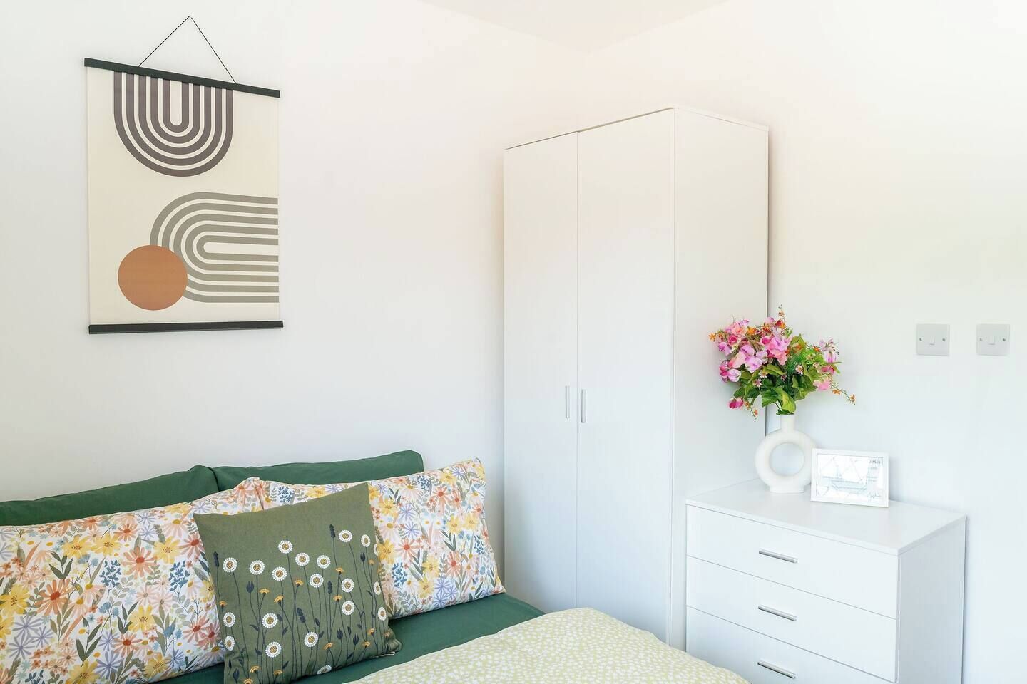 Ein Schlafzimmer mit Bett, Kleiderschrank, Nachttisch mit Blumenvase und einem an der Wand hängenden abstrakten Kunstwerk.