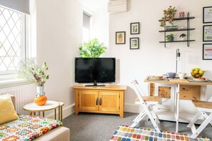 Ein Wohnzimmer mit Fernseher, einem kleinen Tisch mit Obstschale, einem weißen Stuhl, einer Holzkommode und einem farbenfrohen Teppich.