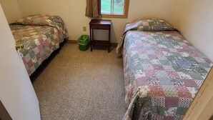 A small room with two beds, a window, a small table, and a trash bin.