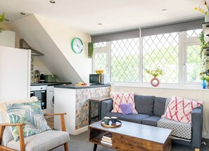 Ein modernes Wohnzimmer mit Sofa, Sessel und Couchtisch. Im Hintergrund ist eine Küche mit Mikrowelle und Spüle zu sehen.