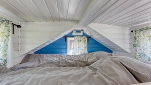 Een slaapkamer met een schuine kap, een bed met grijze beddengoed, en een raam met gordijn.