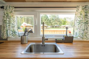 A kitchen with a stainless steel sink, a utensil holder, and a window with a view of a garden and outdoor furniture. - De Glamping - Blue Tiny House | Sleeps 6 (Zeewolde)