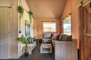 A cozy room with a wooden bench, a sofa, and a small table. There are plants and a window providing natural light. - De Glamping - Duo Lodge | 12p (Zeewolde)