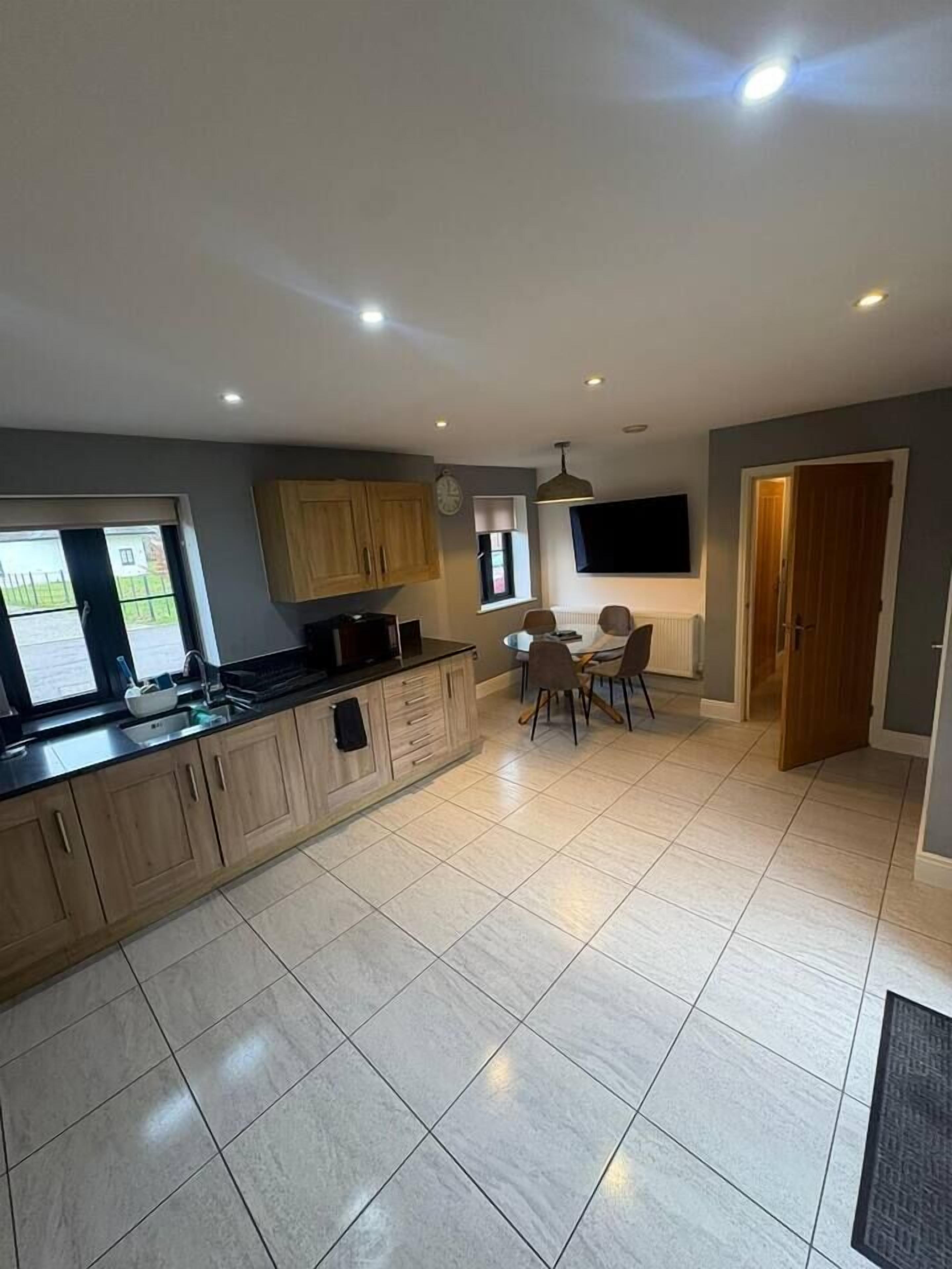 A modern kitchen with a dining area, a flat-screen TV mounted on the wall, and a large window providing natural light.