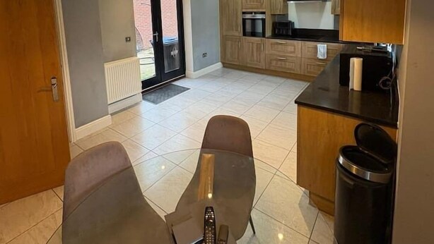 A modern kitchen with wooden cabinets, a dining area with a table and chairs, and a hanging light fixture. - #100 Comfortable 4-Bed House in Rugby (Warwickshire)