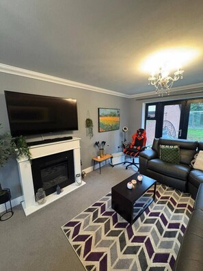 A living room with a fireplace, a flat-screen TV mounted above it, a leather sofa, a patterned rug, and a chandelier. - #100 Comfortable 4-Bed House in Rugby (Warwickshire)