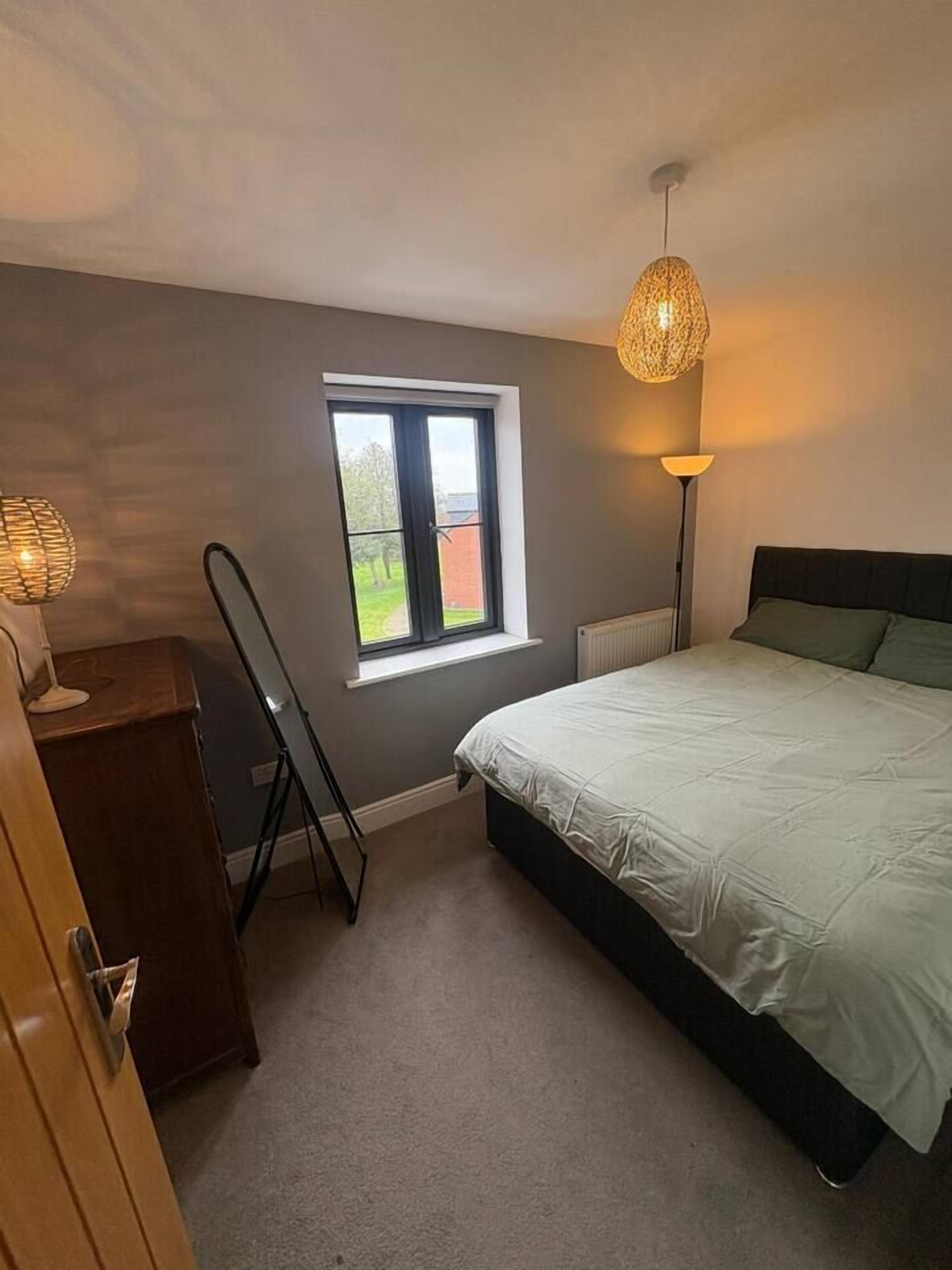A bedroom with a bed, a window, a standing lamp, and a desk.
