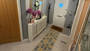 A hallway with a patterned rug, a console table with decorative items, a mirror, and a door with a light switch. - #100 Comfortable 4-Bed House in Rugby (Warwickshire)