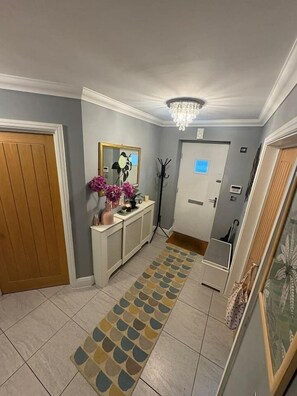 Um corredor com tapete estampado, mesa de apoio com objetos decorativos, espelho e porta com interruptor de luz.