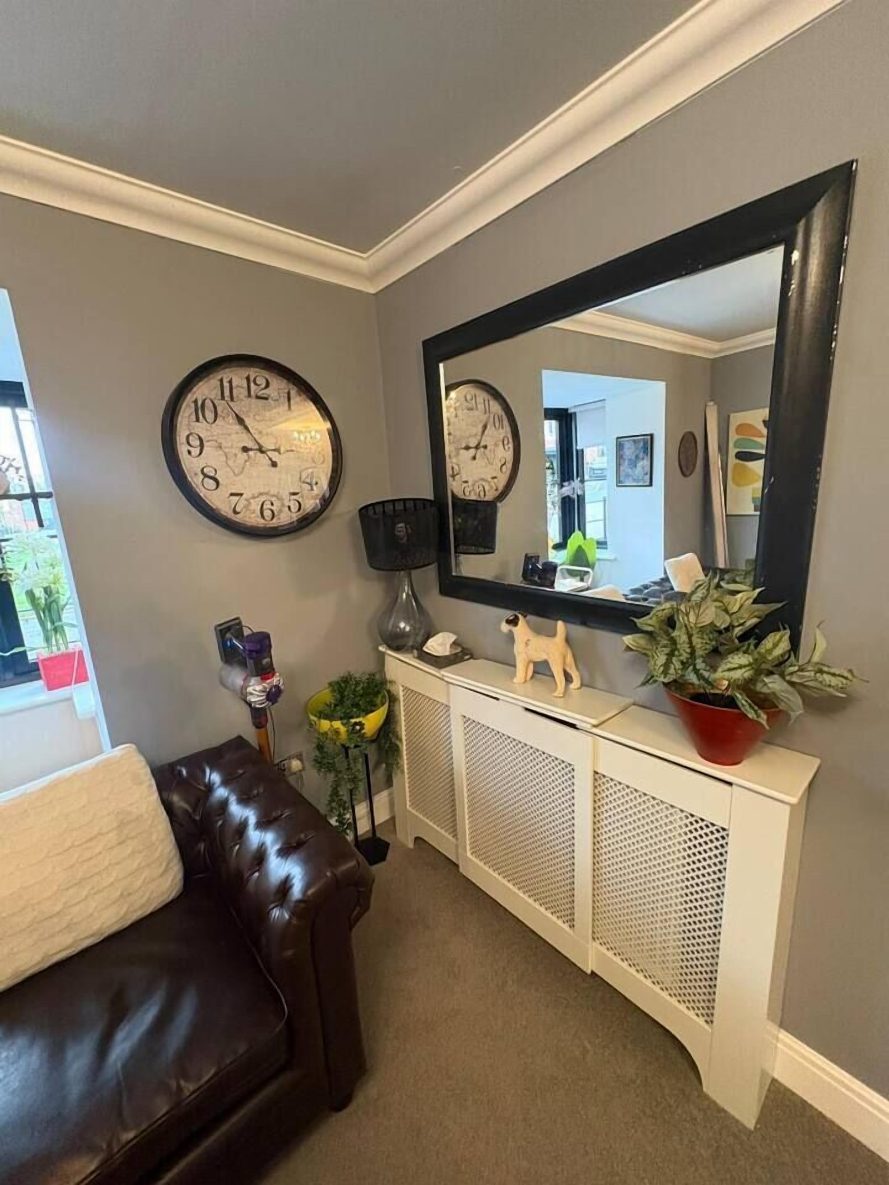 Un salon avec un canapé en cuir marron, une console blanche ornée de plantes, une grande horloge murale et un miroir.