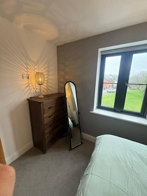 A bedroom with a dresser, a full-length mirror, a bed, and a window with a view of a house and lawn. - #100 Comfortable 4-Bed House in Rugby (Warwickshire)