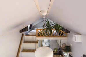 Ett sovrum på entresolvåningen med ett sluttande tak, en säng, en trätrappa, en hängande lampa och en krukväxt.