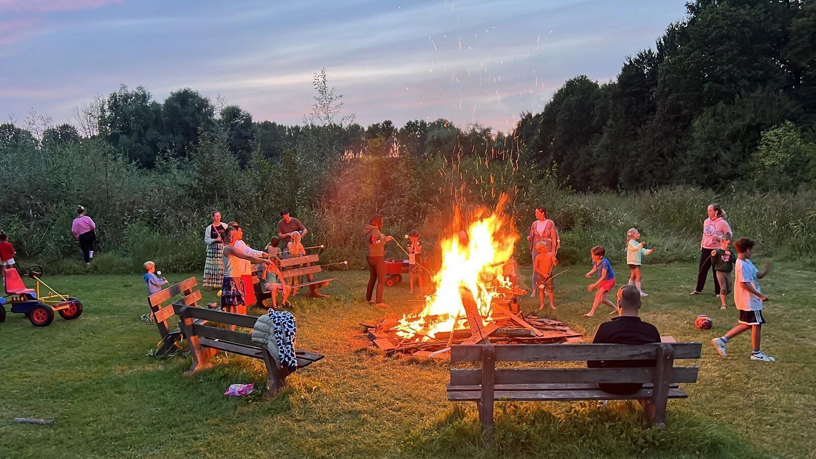 An outdoor gathering with a campfire, people sitting on benches, and a grassy field.