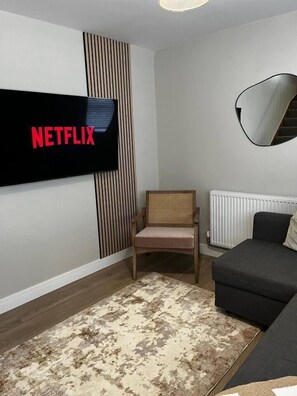 Un salón con un televisor de pantalla plana en la pared, una silla de madera, un sofá negro y un tapete estampado.