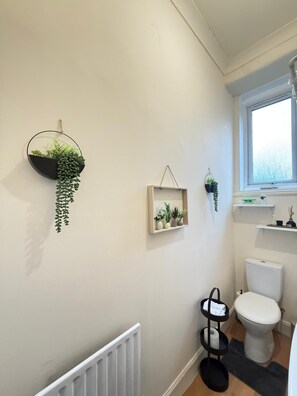 A bathroom with a toilet, a radiator, a window, and wall-mounted shelves with plants. - 2BHK Easter Road, Edinburgh (Edinburgh)