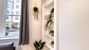 Ein Zimmer mit Fenster, einem grauen Sofa, einer weißen Wand mit Uhr und einem Regal mit Pflanzen.