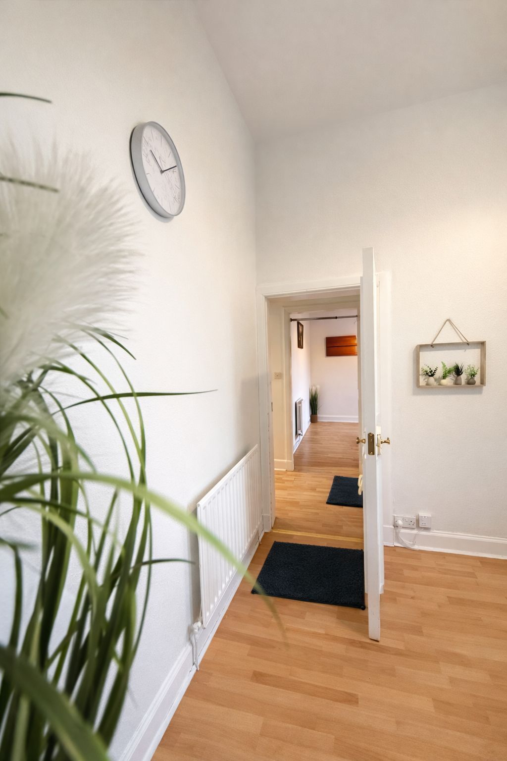 A hallway with a white wall, a clock, a radiator, and a door leading to another room.