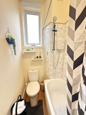 Une petite salle de bain avec des toilettes, une douche, une fenêtre et un sac à serviettes posé sur le sol.