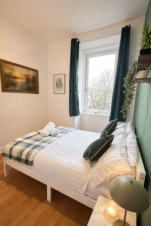 A neatly made bed with a checkered blanket, a bedside lamp, and a window with curtains. - 2BHK Easter Road, Edinburgh (Edinburgh)