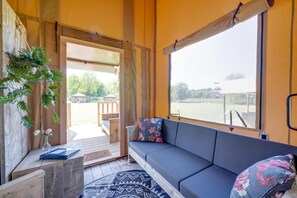 A room with a wooden floor, a sofa with floral cushions, a small table with a vase, and a view of a deck and outdoor area. - De Glamping - Sunny Lodge | Sleeps 6 (Zeewolde)