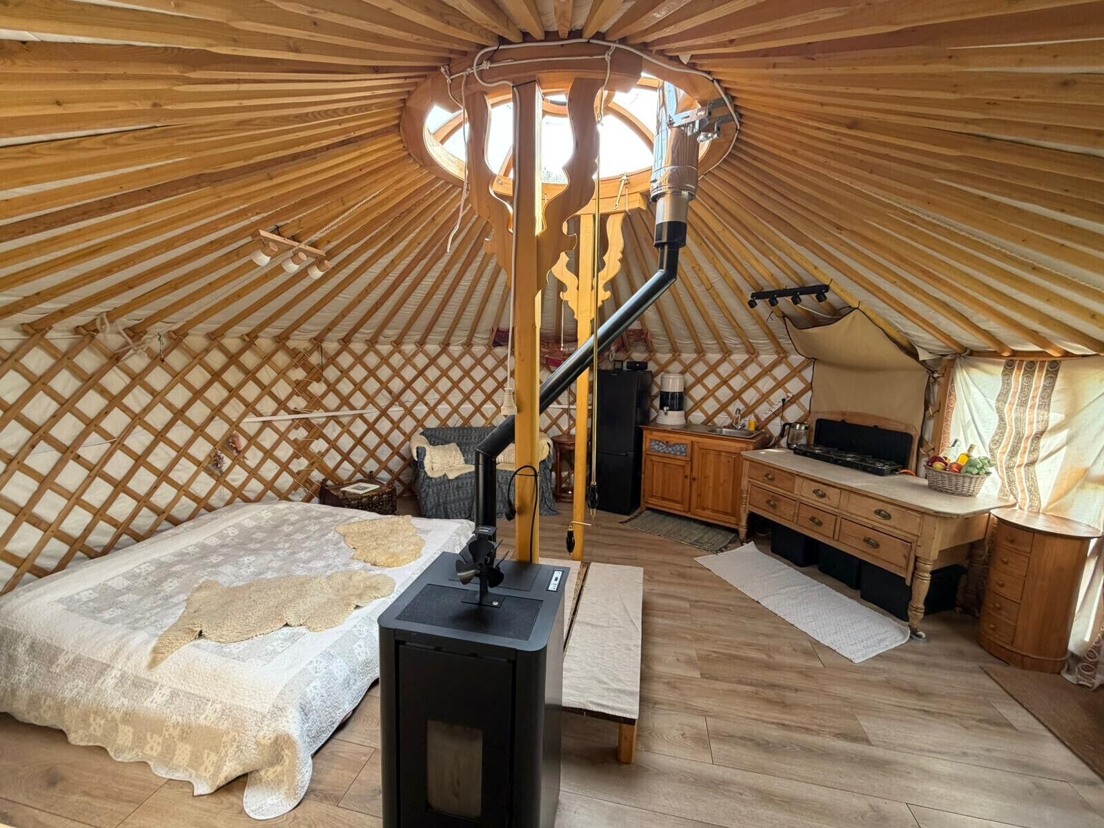 A yurt interior with a central light fixture, a bed with patterned bedding, a small table, and a kitchenette area.