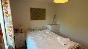 Ein Einzelbett mit weißer Daunendecke, ein Nachttisch mit Lampe, eine kleine Zimmerpflanze und ein Wandbild.