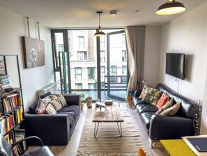 A modern living room with a black leather sofa, a coffee table, a bookshelf, and a large window offering a view of buildings. - #177 Stylish Finsbury Park Retreat - Sleeps 2 (Greater London)