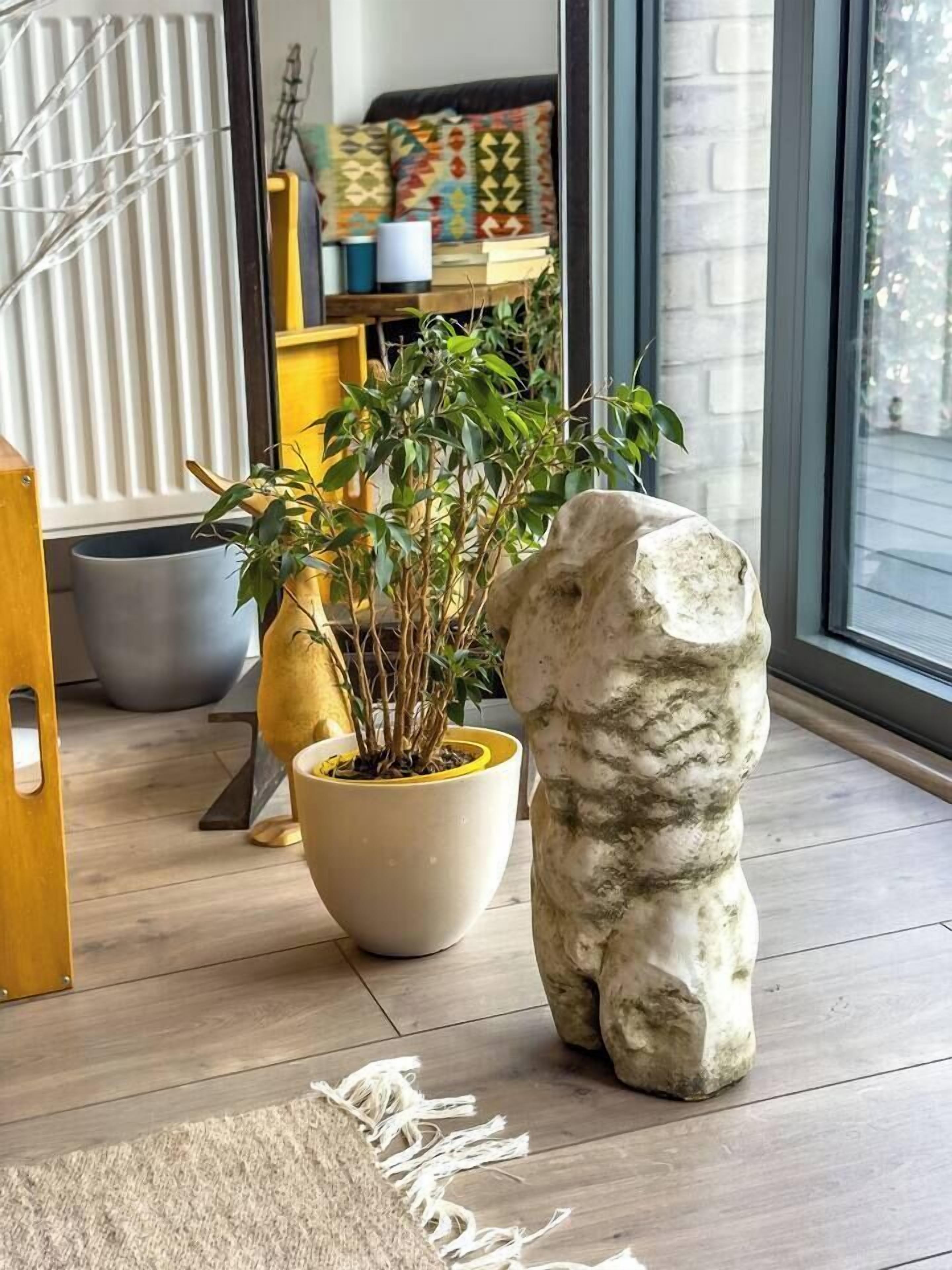 Una escultura de piedra, una planta en maceta y una silla de madera en una habitación con un ventanal grande y un cojín estampado en un estante.