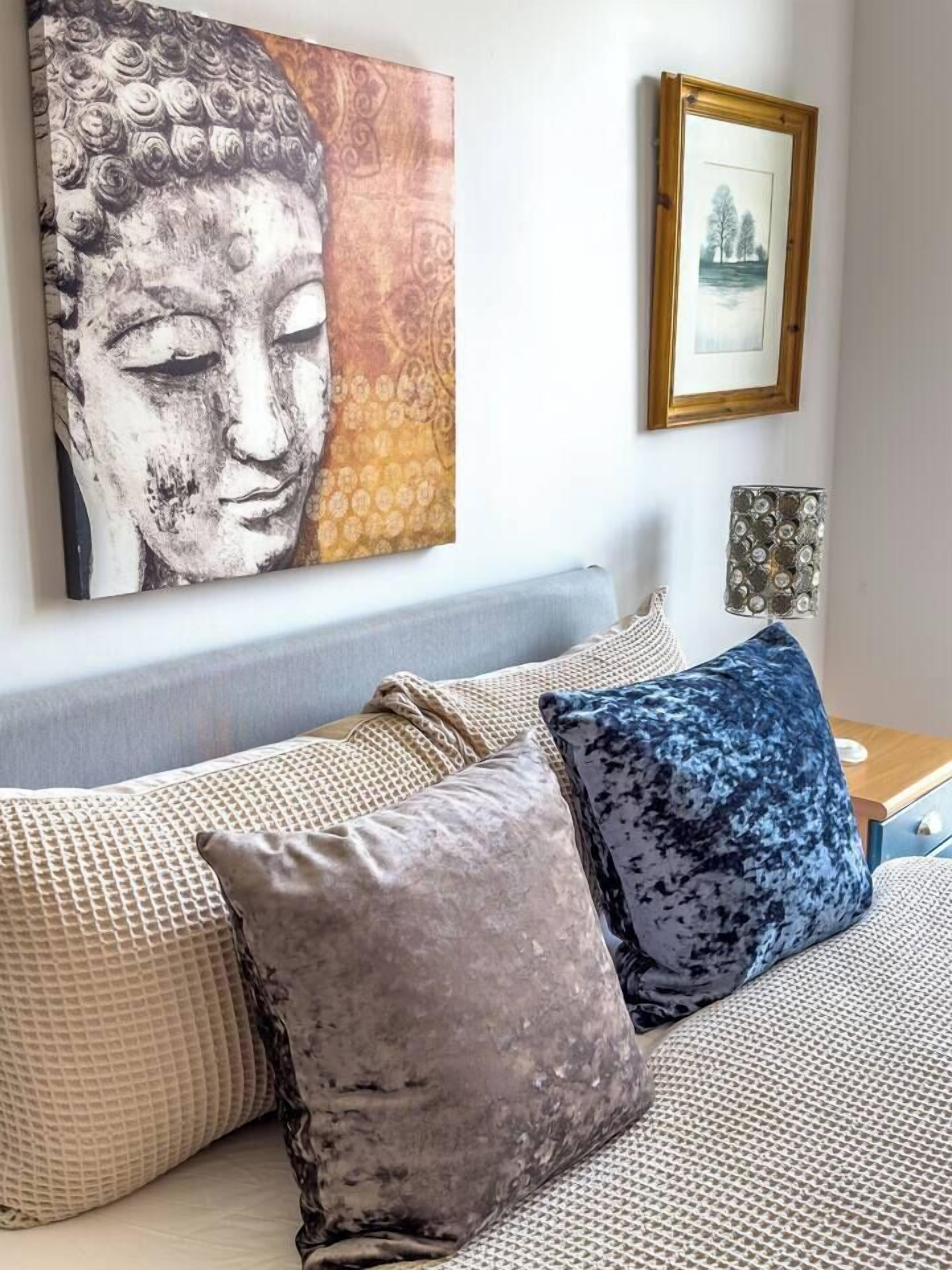 Un dormitorio con cama, almohadas, un cuadro en la pared y una mesita de noche con lámpara.