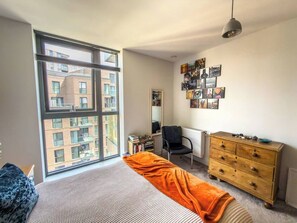 Ein Schlafzimmer mit großem Fenster, einem Bett, einem Schreibtisch, einem Sessel, einer Kommode und Wandbildern.