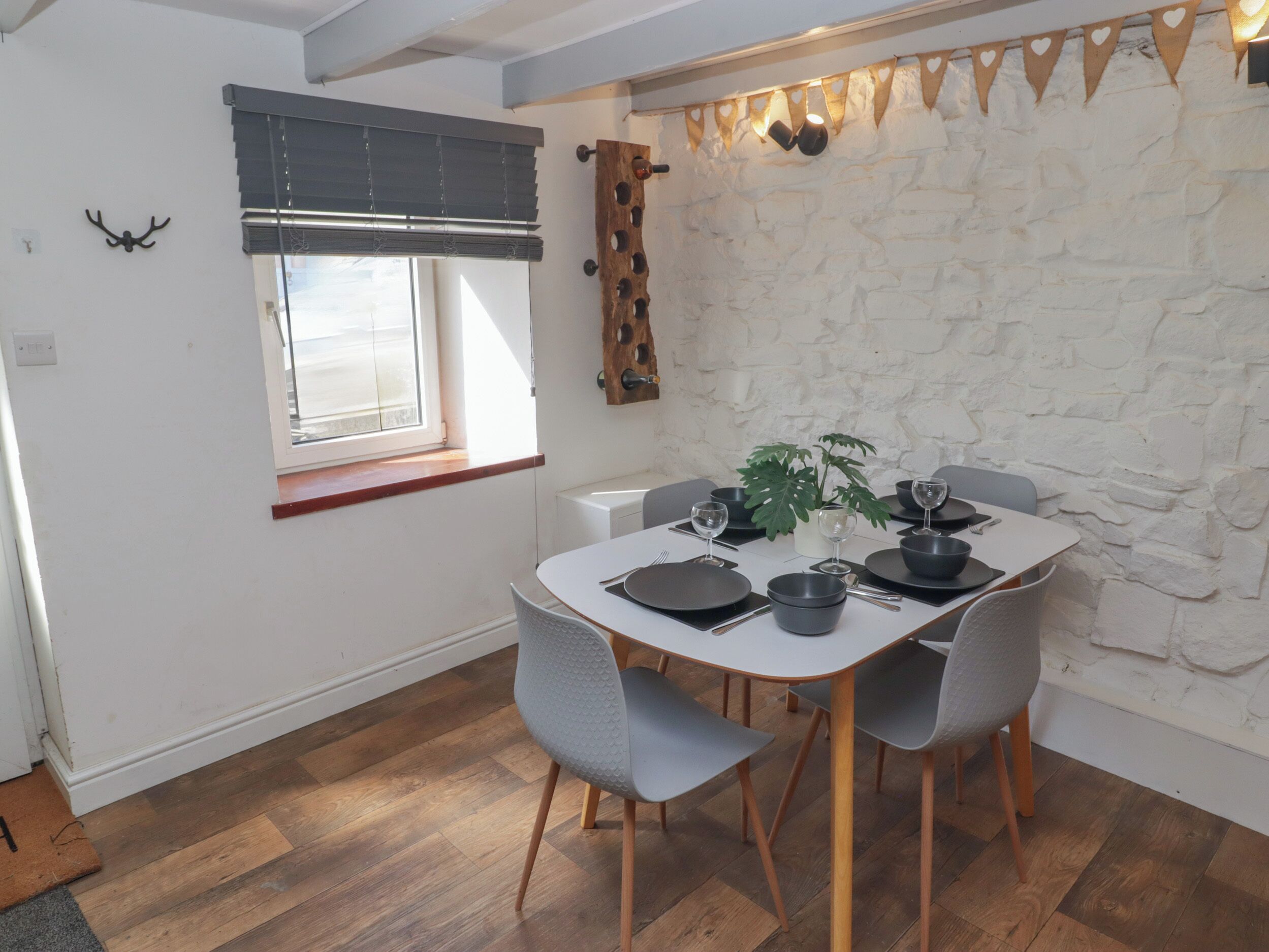 A dining area with a white table set for four, surrounded by grey chairs, a potted plant, and a stone wall.