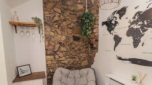 Ein gemütliches Zimmer mit Steinwand, grau gepolstertem Sessel, einer Weltkarte und einem Regal mit einer Flasche und Gläsern.