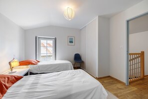 A bedroom with two beds, a desk with a lamp, a chair, a window, and a door.