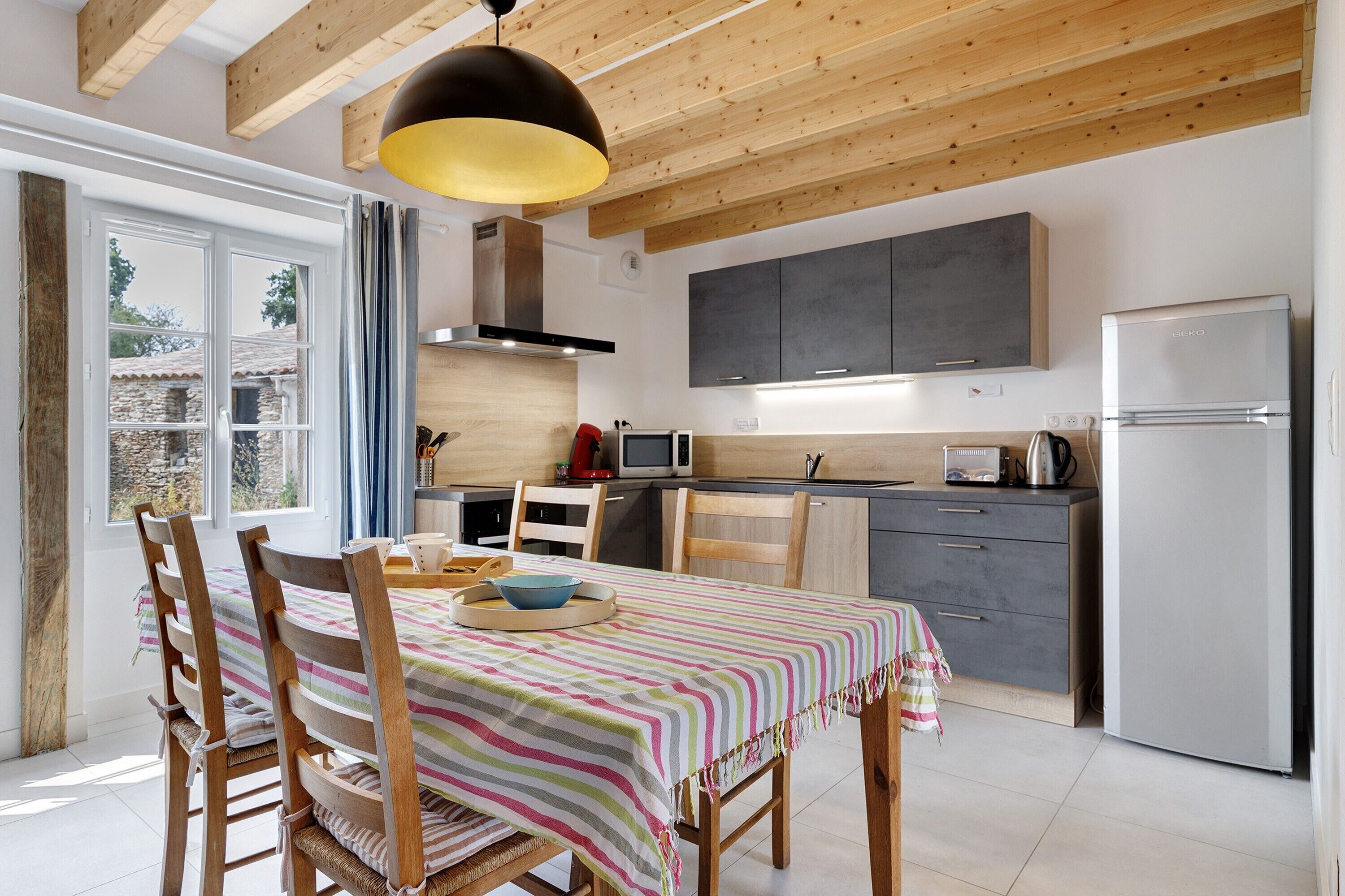A modern kitchen with wooden beams, a dining table with striped tablecloth, and a large window.