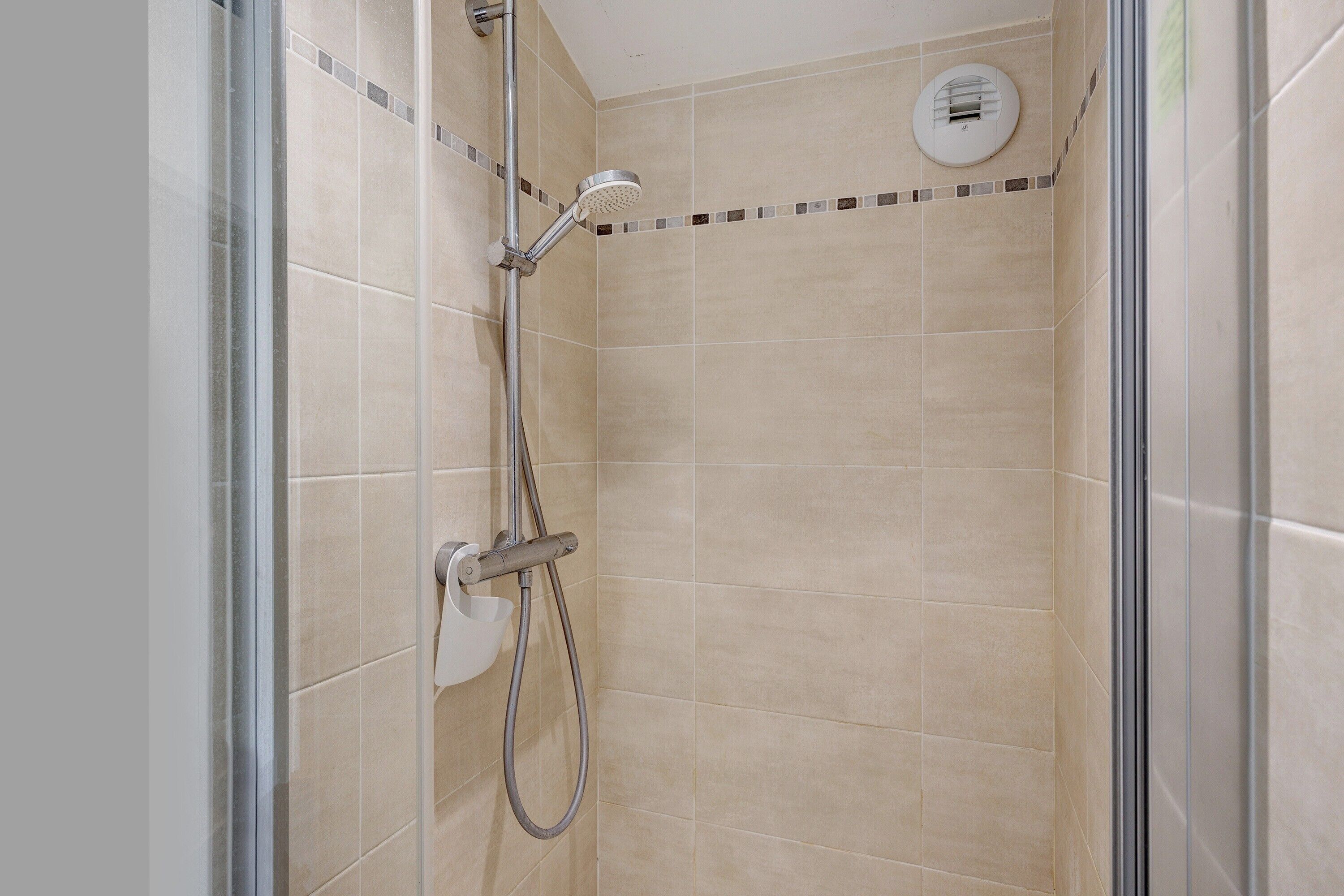 Une cabine de douche fermée par des parois en verre, avec des carreaux de couleur beige, une douchette à main et un panneau de commande fixé au mur.