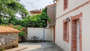 Um pátio com muro de pedra, uma mesa com cadeiras e um edifício com tijolos vermelhos e paredes brancas.