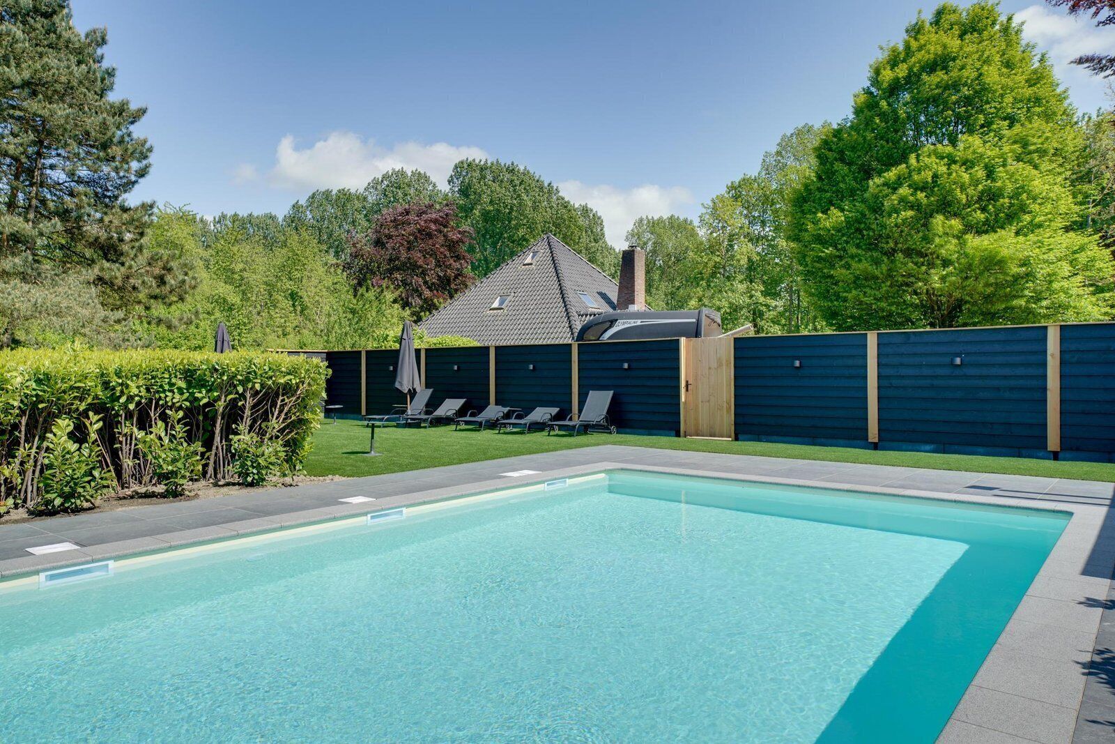 Ein rechteckiger Pool mit Fliesenrand, umgeben von einem dunklen Zaun, und ein Gebäude mit geneigtem Dach im Hintergrund.